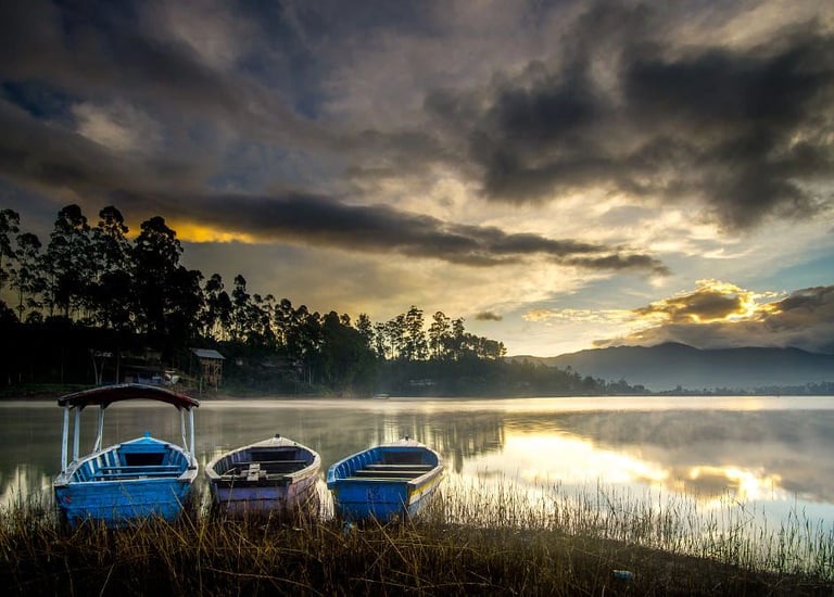 Tiga perahu bersandar di tepi Danau Situ Cileunca saat matahari terbit, dengan kabut tipis dan pantulan langit di permukaan a