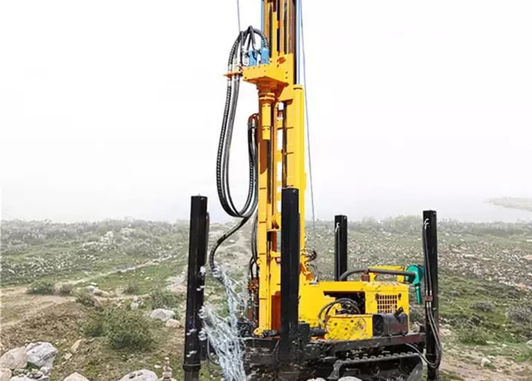 Yellow crawler-mounted hydraulic water well drilling rig operating on a rocky construction site.