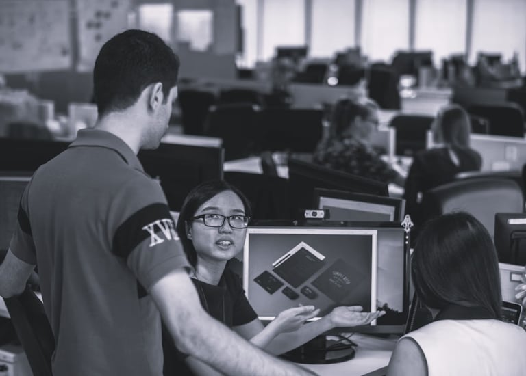 three colleagues, two sat by a computer with another stood with them discussing options for a projec
