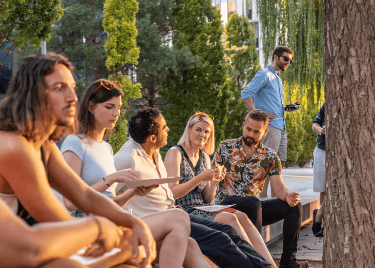 A croup of coworkers sitting on stairs at an event talking. Green trees in the background, sunlight hitting their faces.