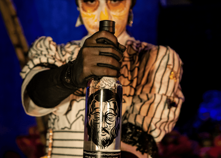 A person in Catrina makeup holding a bottle of Mezcal Judas artisanal Mexican spirit.
