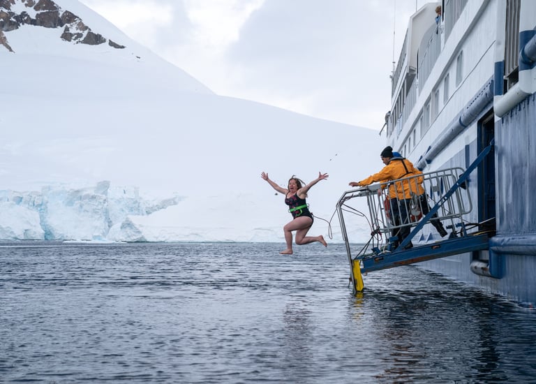A tourist does a polar plunge from a cruise ship into icy Antarctic waters near snowy mountains.