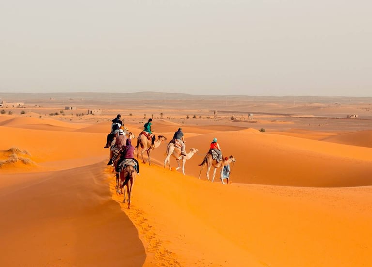 Tourists riding camels in a caravan across the orange sand dunes of the Sahara Desert.