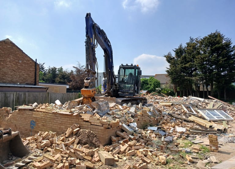 Heavy excavator clearing brick rubble and debris at a residential demolition site.