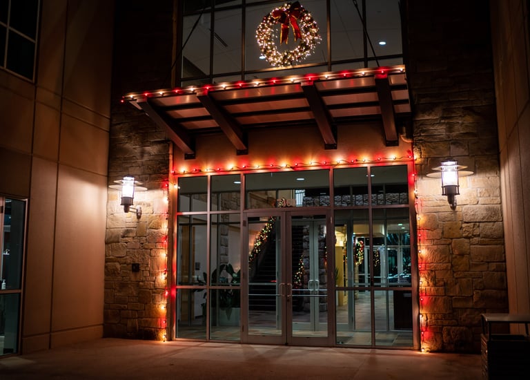 Office building glass entrance decorated with red Christmas lights and a lit holiday wreath at night.