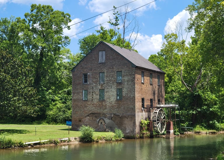 A historic brick watermill with a large wooden wheel sits beside a calm river in a lush green forest.