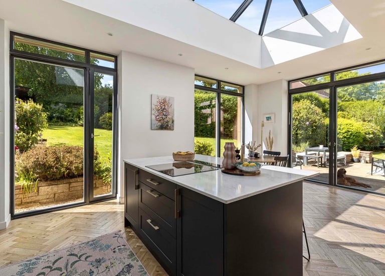 Modern open-plan kitchen featuring a dark island, skylight, and large glass doors opening to a lush garden.