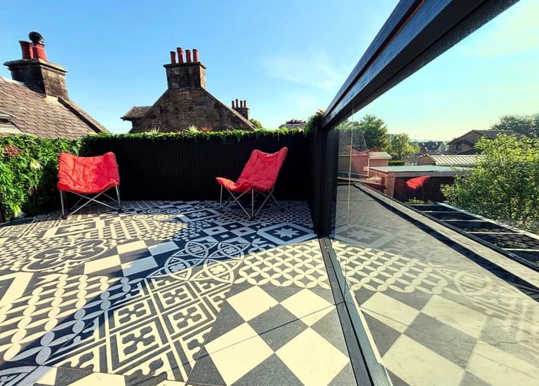 Modern rooftop terrace with patterned tile flooring, red lounge chairs, and privacy greenery overlooking the city.