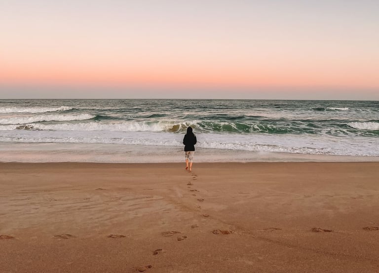Uma pessoa andando em direção ao mar, de costas, ao por do sol.