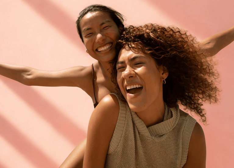 Twee lachende vrouwen, één draagt de ander op de rug tegen een roze achtergrond.