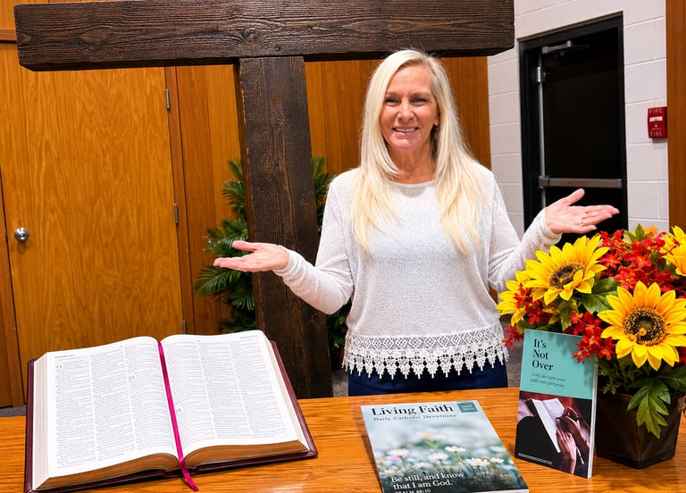 Myra Lee standing by a wooden cross with an open Bible and Daily Catholic Devotions book.