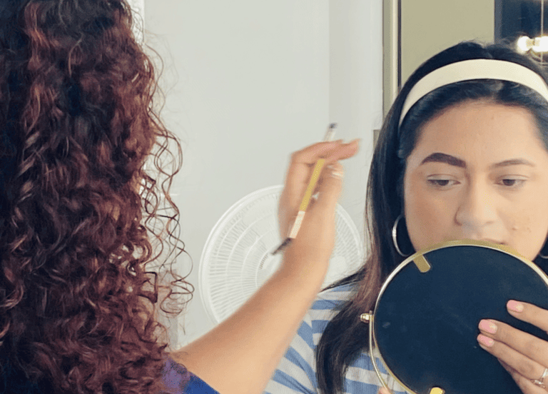 Maquilladora profesional aplica lápiz de cejas a una mujer que sostiene un espejo en un estudio.