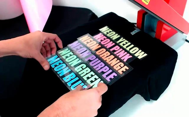 Applying neon heat transfer vinyl labels to a black t-shirt using a professional heat press machine.