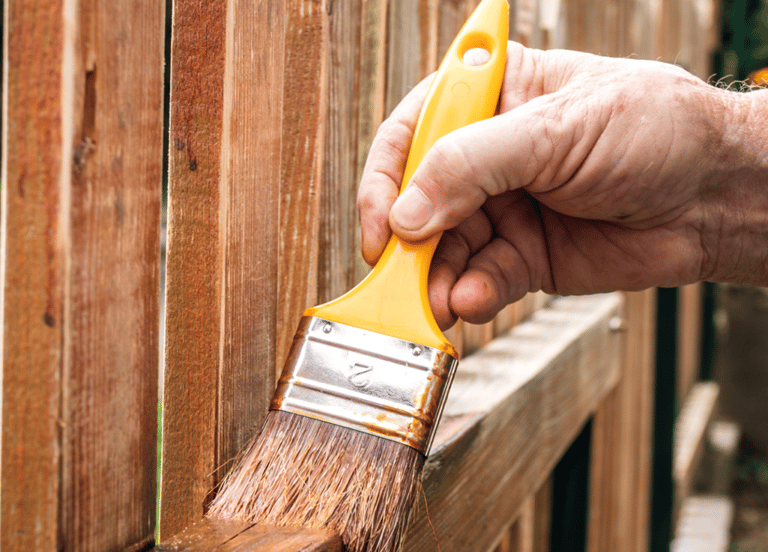 Close-up of a landscaper applying protective wood sealer on a cedar fence post with a brush.