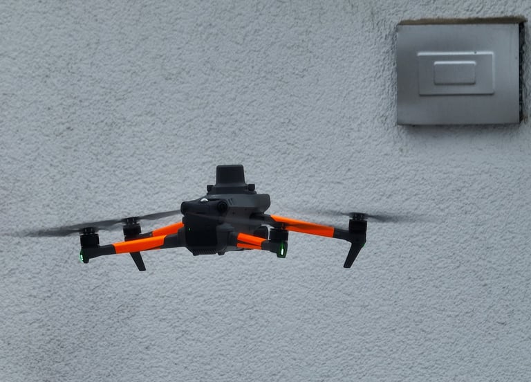 Professional orange and black quadcopter drone hovering near a grey textured building wall.