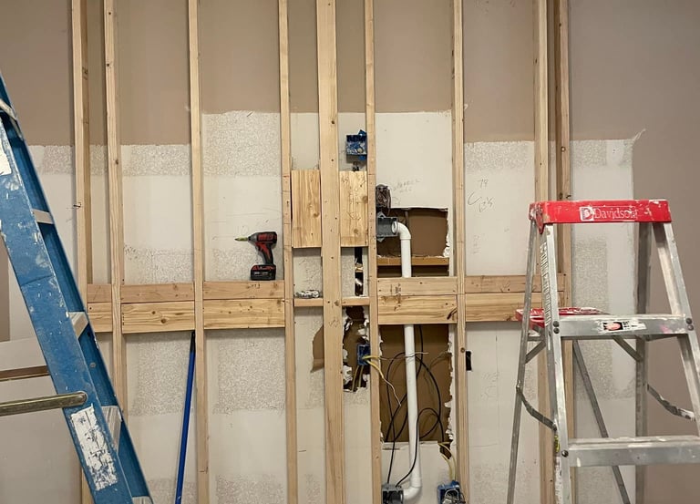 Indoor wall framing renovation with wooden studs, electrical wiring, and plumbing pipes visible between two ladders.