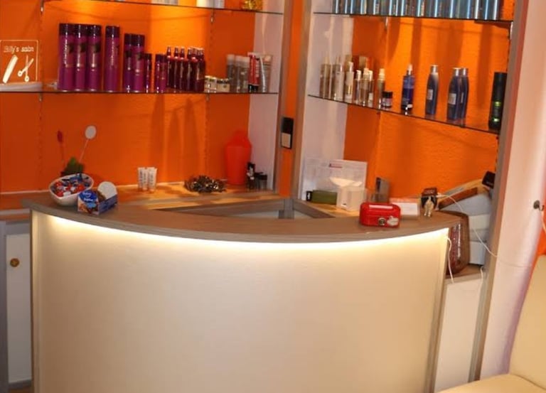 A modern hair salon reception desk with orange walls and glass shelves displaying professional beauty products.