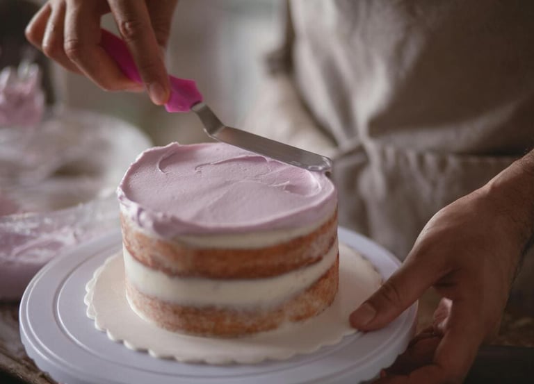 Baker using an offset spatula to spread purple frosting on a small layered sponge cake.