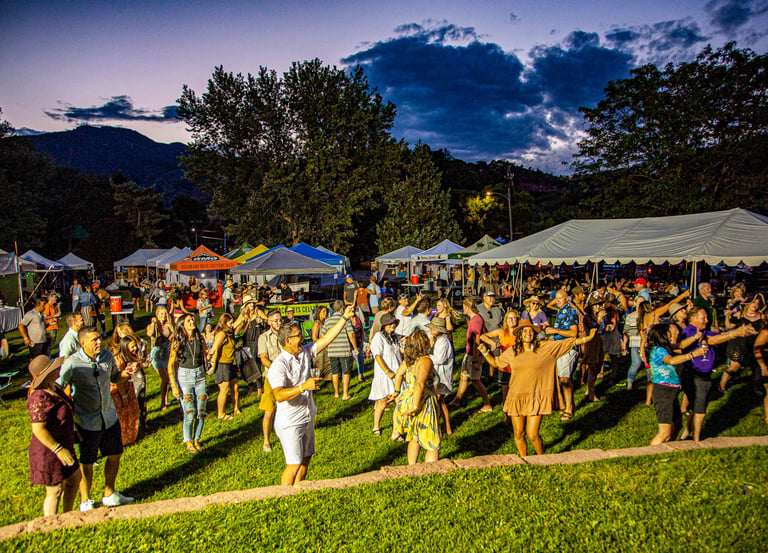 Crowd dancing at an outdoor summer music festival with event tents and mountains at sunset.