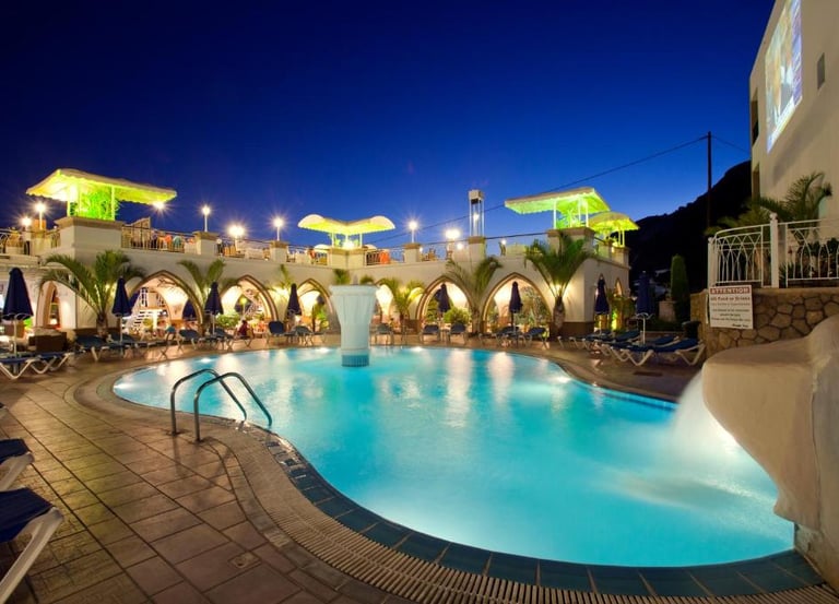 Illuminated outdoor resort swimming pool at night with lounge chairs and palm trees.