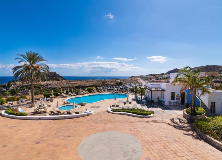 Luxury resort pool area overlooking the ocean in Rhodes, Greece with palm trees and sun loungers.