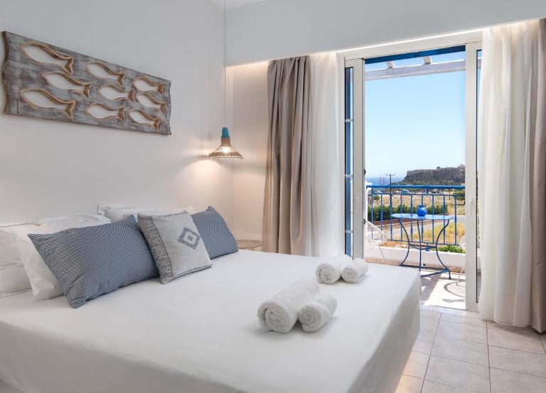 Modern Mediterranean hotel room with white bed linens, blue pillows, and a balcony overlooking the sea.