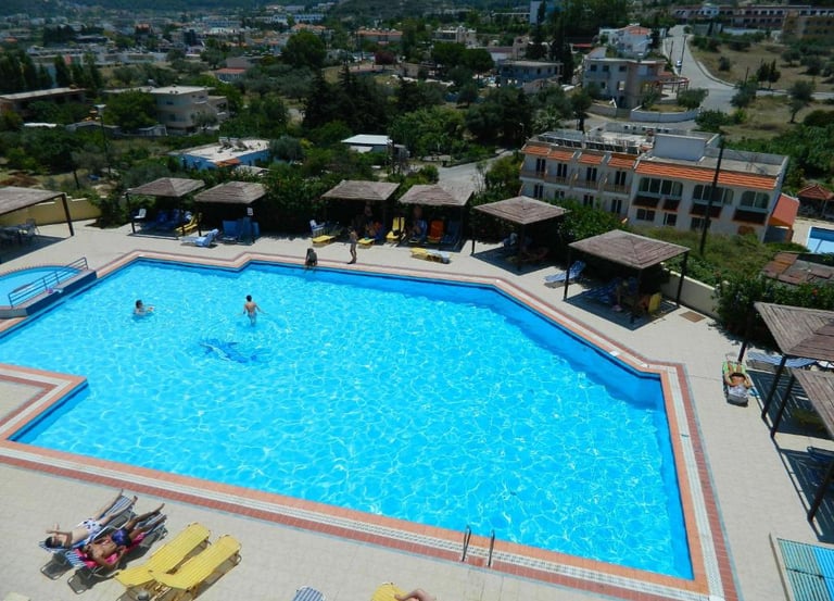 Aerial view of a bright blue swimming pool at a Mediterranean hotel resort with sun loungers and cabanas.
