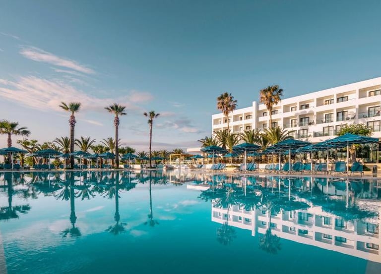 Luxury beach resort with a large swimming pool, palm trees, and blue umbrellas under a clear sky.