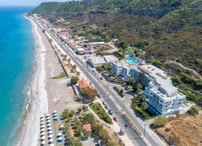 Aerial view of a coastal resort and sandy beach in Rhodes, Greece, with turquoise sea and green hills.