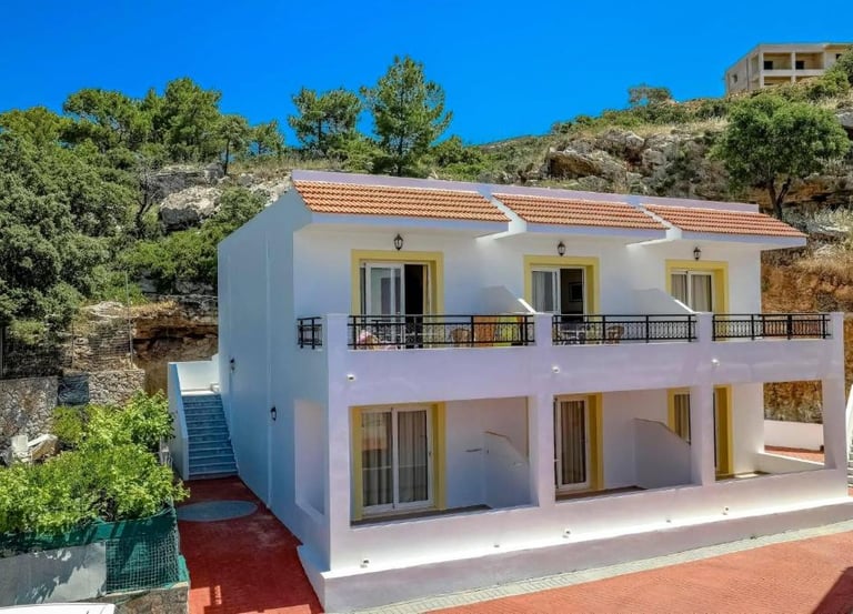Two-story white Mediterranean villa with balconies nestled against a rocky hillside under a clear blue sky.