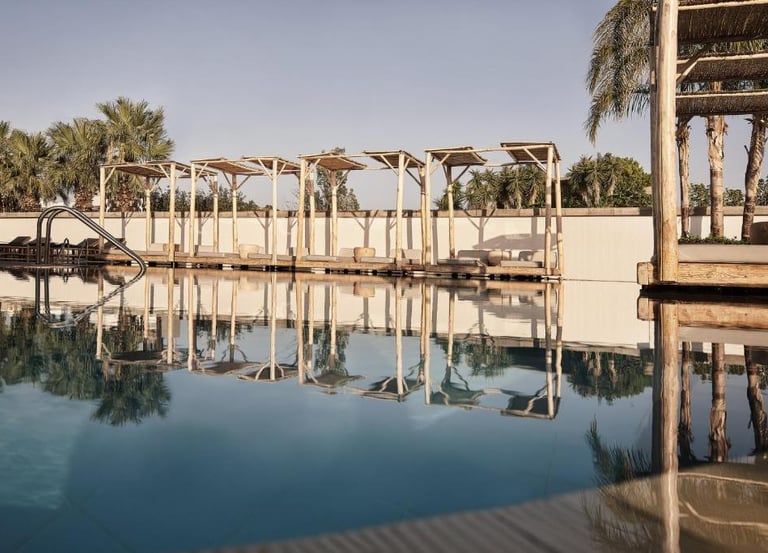 Luxury resort swimming pool with wooden cabanas and palm tree reflections on calm water.