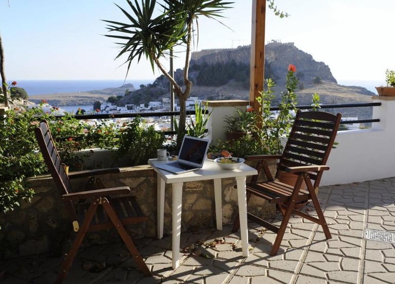 Laptop on a terrace table with wooden chairs overlooking Lindos Acropolis and the sea.