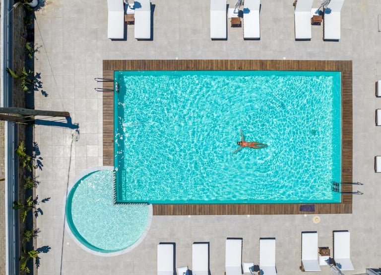 Aerial view of a woman floating in a luxury resort swimming pool with a sun deck and white lounge chairs.