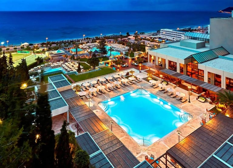 Luxury beach resort with illuminated swimming pools and tennis courts at dusk overlooking the ocean.