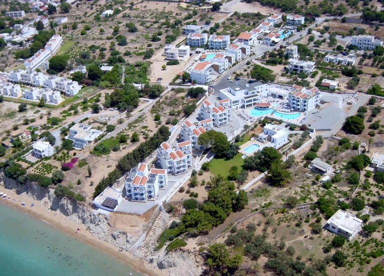 Aerial view of a coastal Greek island resort with white villas and swimming pools near the beach.