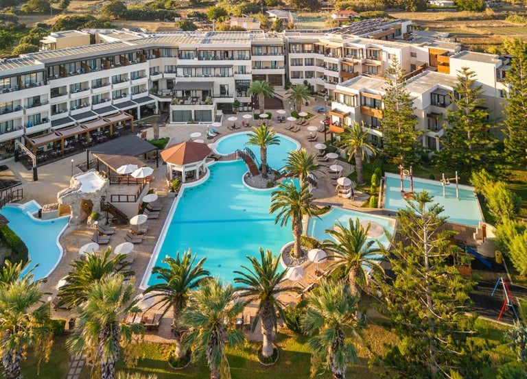 Aerial view of a luxury Greek island resort featuring turquoise swimming pools, palm trees, and modern hotel suites.
