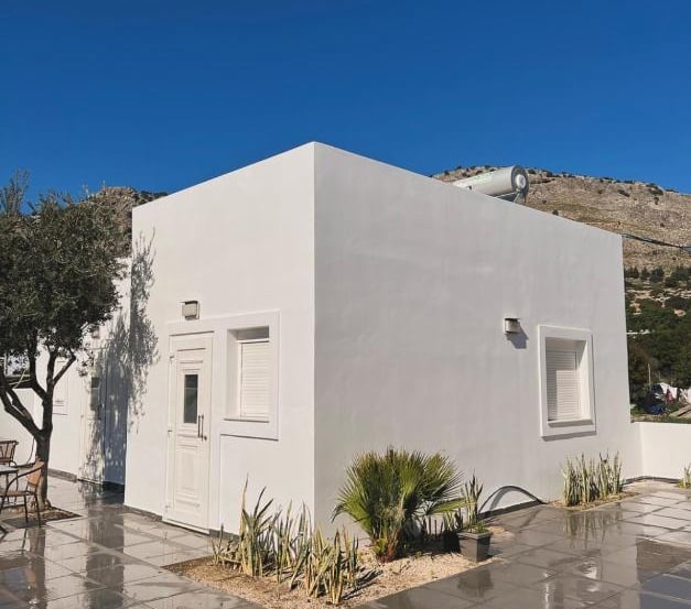 Modern white minimalist villa with a tiled terrace and olive trees under a clear blue sky.