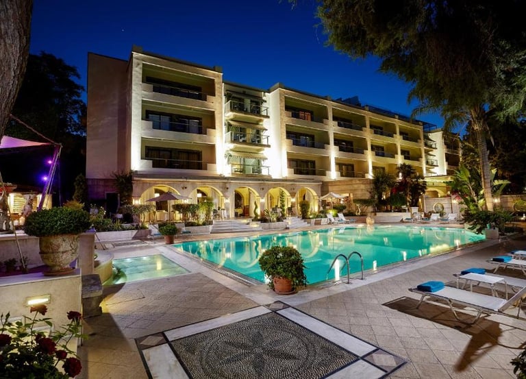 Luxury hotel exterior at night with illuminated swimming pool and sun loungers.