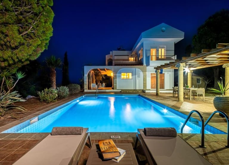 Luxury villa with an illuminated swimming pool and lounge chairs at night.
