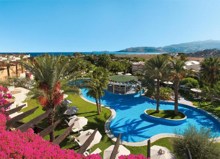 Luxury beach resort with a large blue swimming pool, palm trees, and ocean views in a tropical landscape.