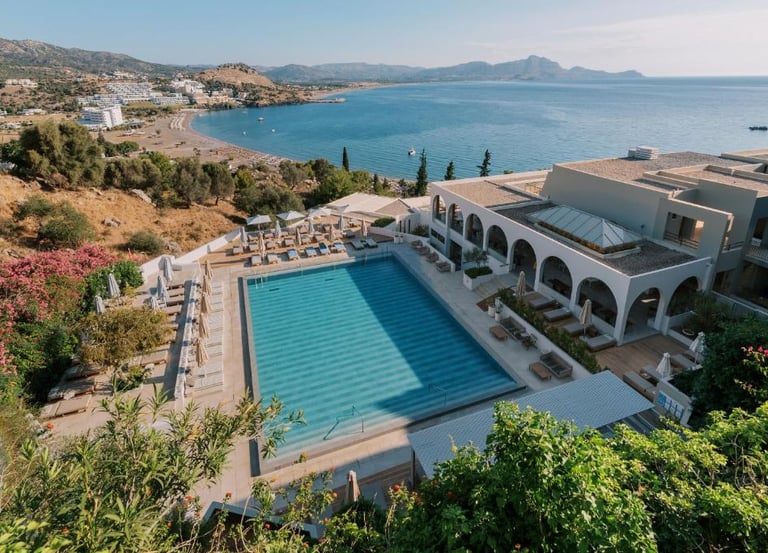Luxury resort swimming pool overlooking a Mediterranean bay and beach at sunset.