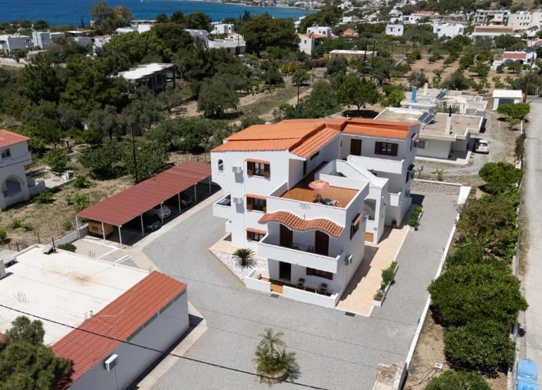 Aerial view of a white multi-story holiday villa with orange roof tiles in a coastal Greek village.