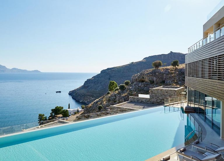 Luxury infinity pool at a coastal resort overlooking the Aegean Sea and rocky cliffs.