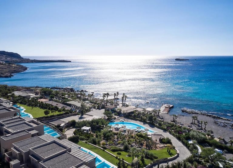 Aerial view of a luxury seaside resort with pools and villas overlooking the blue Aegean Sea.