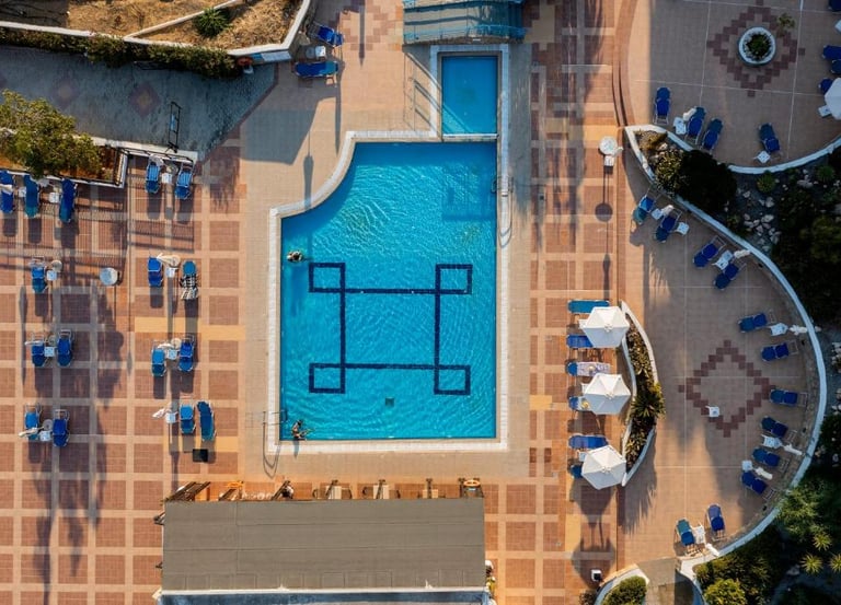 Aerial view of a luxury hotel swimming pool with blue lounge chairs and sun umbrellas on a tiled patio.