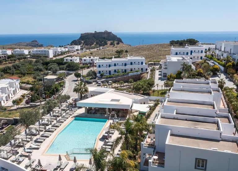 Aerial view of a luxury Greek island resort with a blue swimming pool and white villas near the sea.