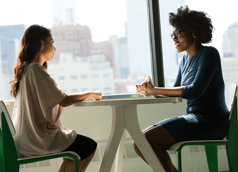 Two professional women sit talking at a workspace table in an office environment.