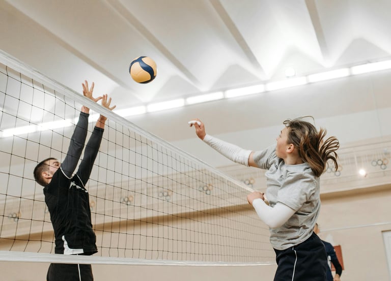 A volleyball player in a gray shirt jumps to spike the ball while an opponent in black reaches up to