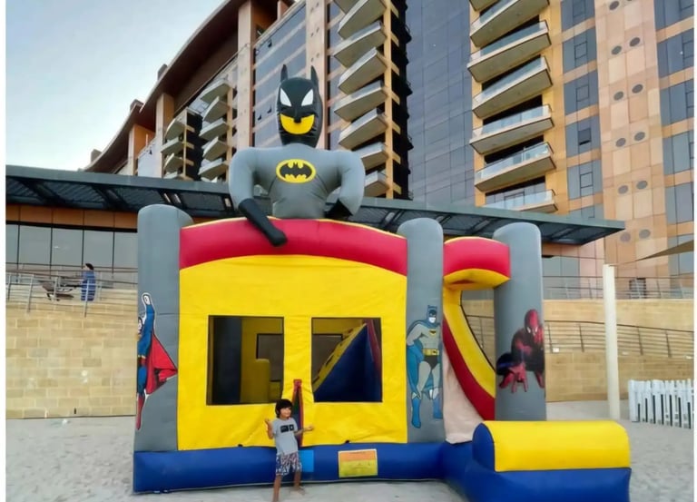 Superhero themed inflatable bounce house with Batman and a slide on a sandy beach.