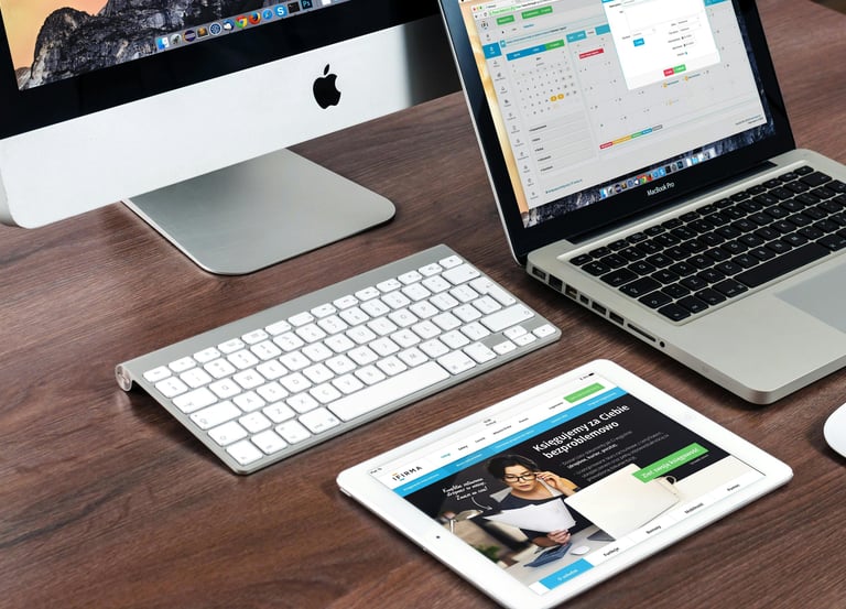 A modern office workspace with an Apple iMac, MacBook laptop, wireless keyboard, and iPad on a wooden desk.